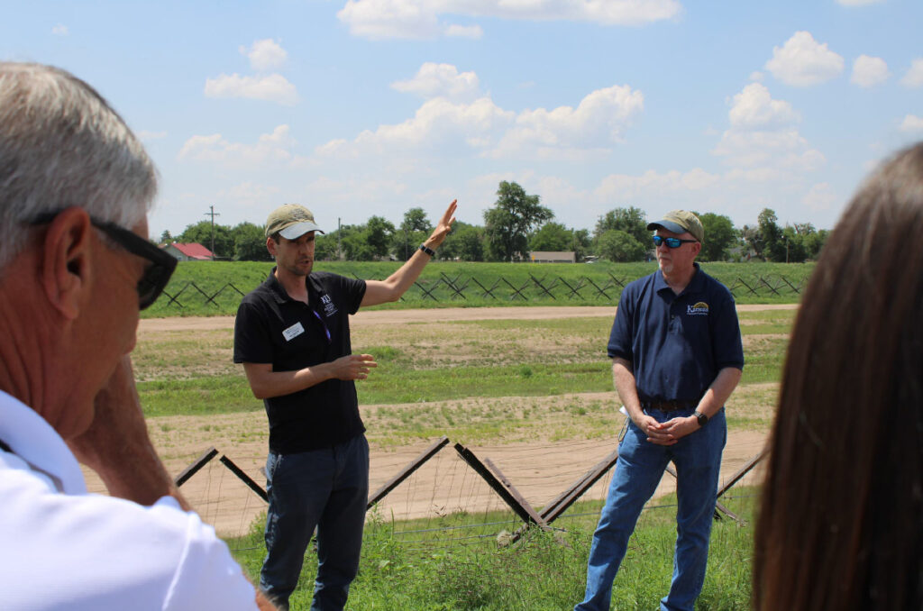 Two people stand in grass near a fence, one speaking and gesturing to the dry riverbed behind him, as others in the foreground listen.