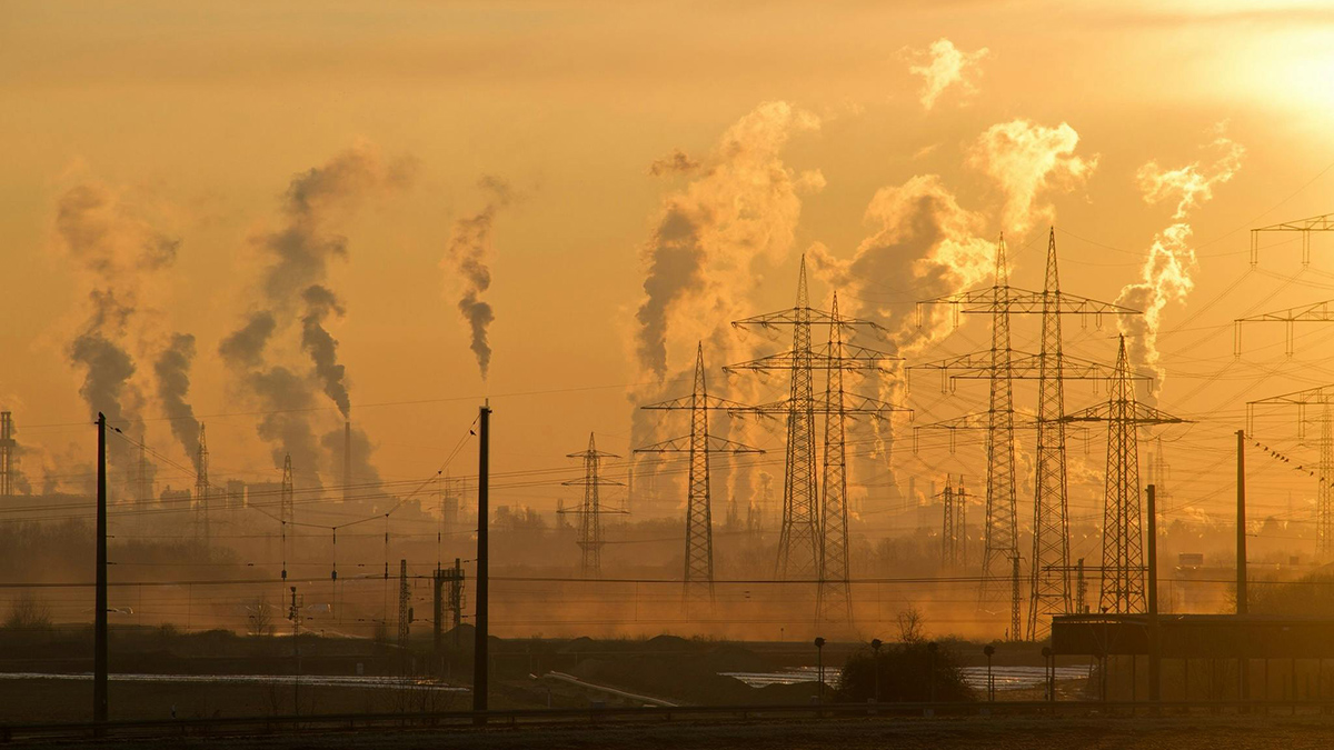 Emissions plumes rise from smokestacks in the distance, behind electrical towers silhouetted against a hazy golden sky.