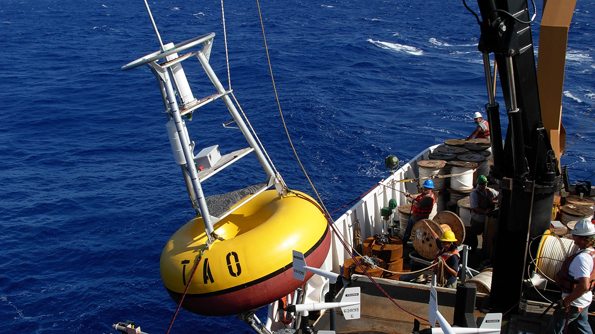A buoy with a red and yellow bottom and a white top, labeled “TAO,” is lowered into the ocean from a ship.
