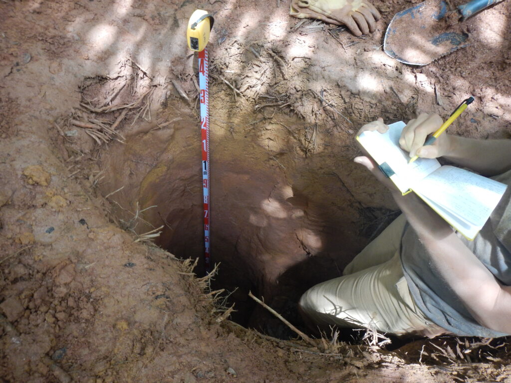 A scientist stands in a deep hole made in reddish soil. A tape measure stands in the hole, and different bands of soil colors and textures can be seen.