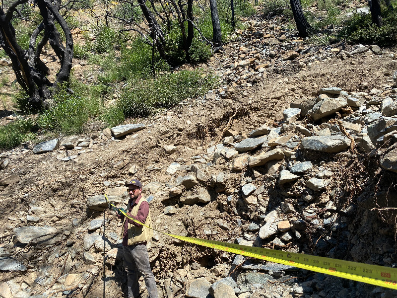 A researcher stands in a channel carved by a debris flow on a rocky hillslope, holding an outstretched measuring tape across the channel.