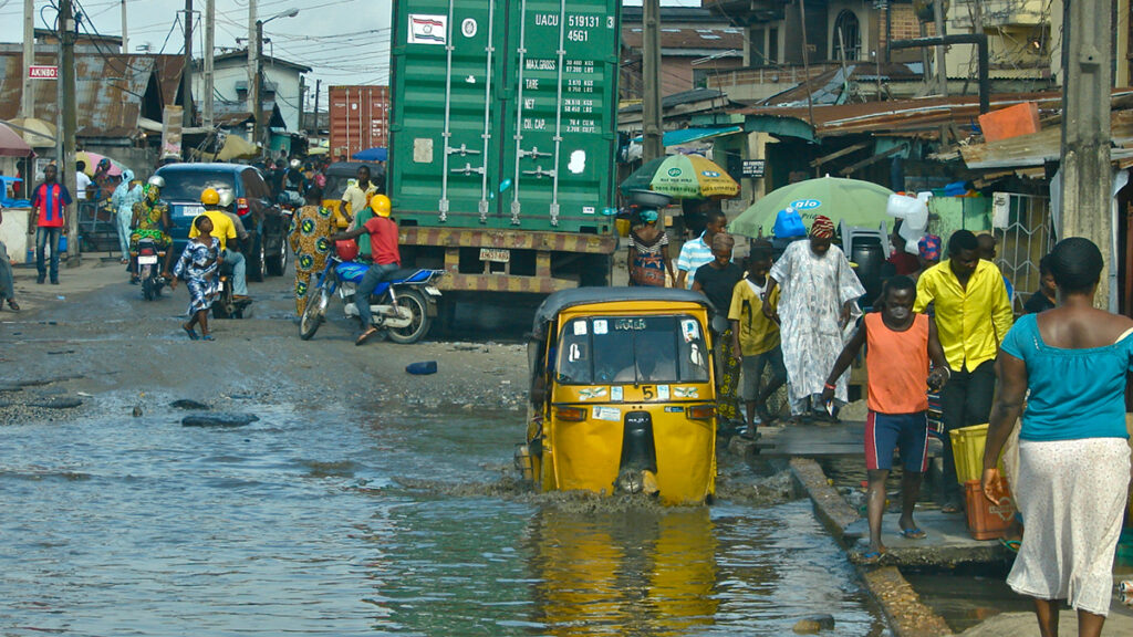 On a flooded street in Lagos, a yellow vehicle’s wheels are submerged in water, and people walk around the water on a sidewalk.