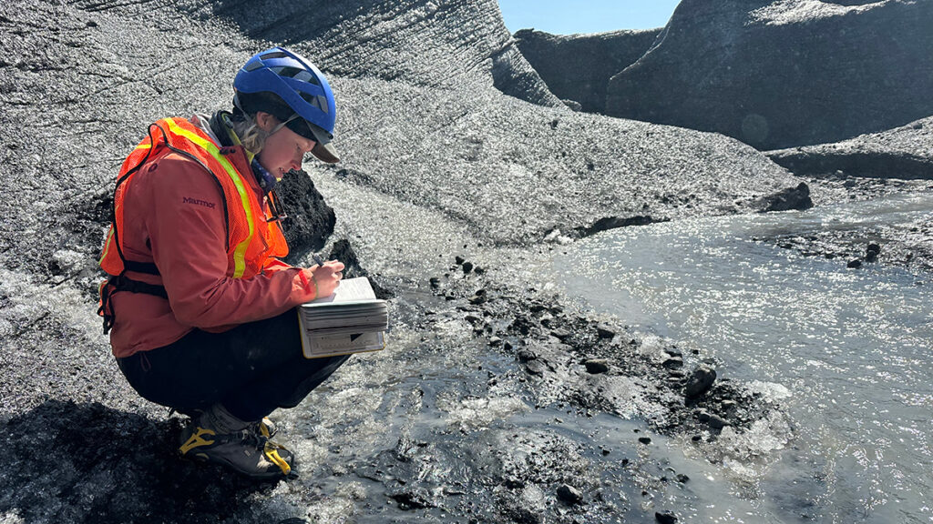 A scientist wearing a safety vest and a blue hard hat squats alongside a stream, taking notes in a notebook. The stream cuts through a glacier covered in dark gray sediment.