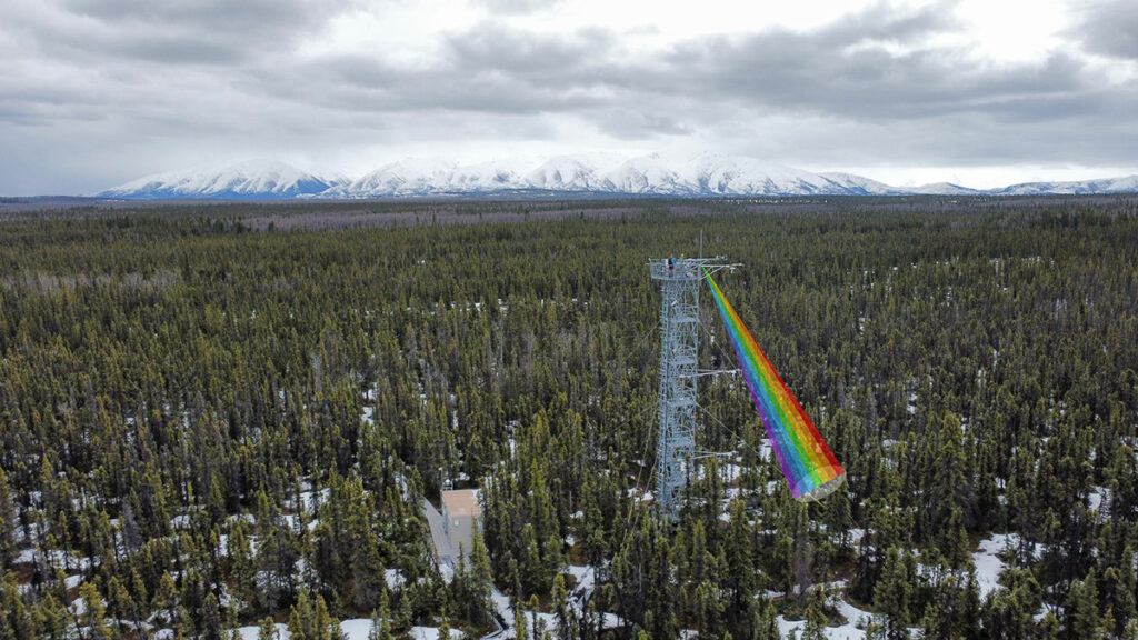 Aerial view of a tall metal frame tower towering over a forest under a cloudy sky and with snow-covered mountains in the distance. An illustrated rainbow-colored beam from the tower to the ground is superimposed on the photo.