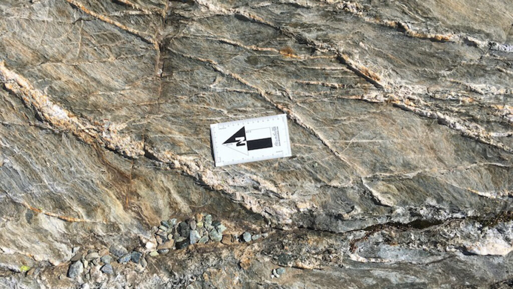 A sheet of gray and brown rock with several large veins running across it diagonally. A flat, white, rectangular measurement device is in the center of the frame.