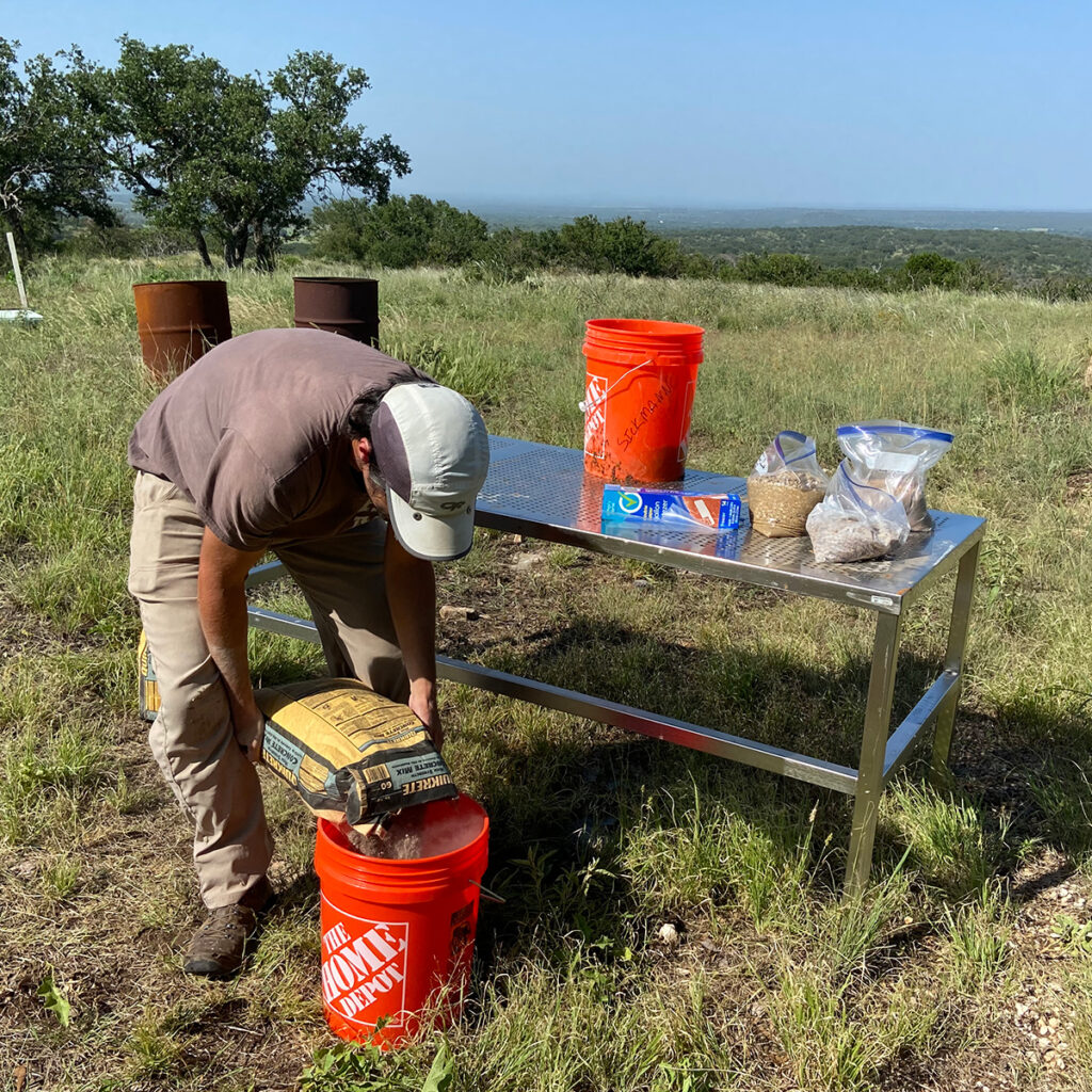 Zachary Sickmann pours cement mix into an orange Home Depot bucket in a green field. He stands beside stainless steel table upon which sit several gallon-sized zip-top plastic bags containing earth and rock, a blue box of more zip-top bags, and a second orange Home Depot bucket.