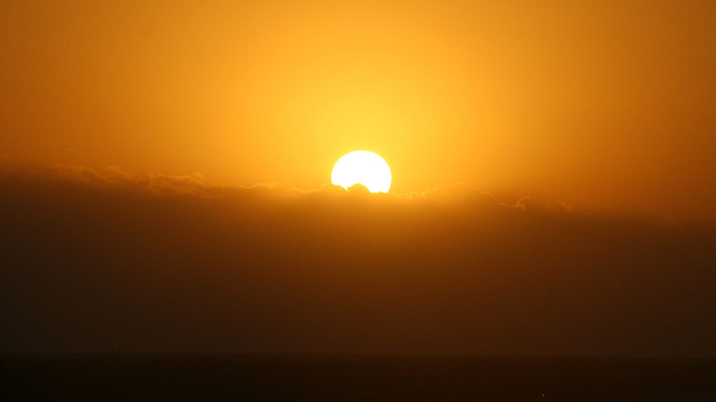 An orange Sun rises over a bank of clouds.
