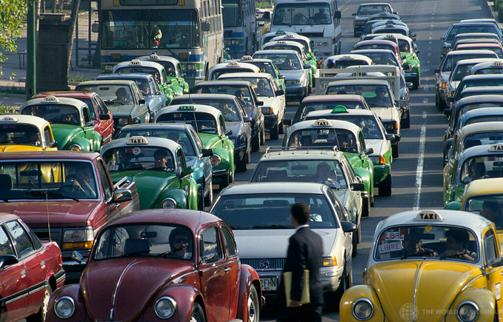 Cars sit in multiple lanes of traffic on a congested road in Mexico. A man in a business suit with a yellow object under his arm is crossing the road in front of the vehicles with his back to the camera.
