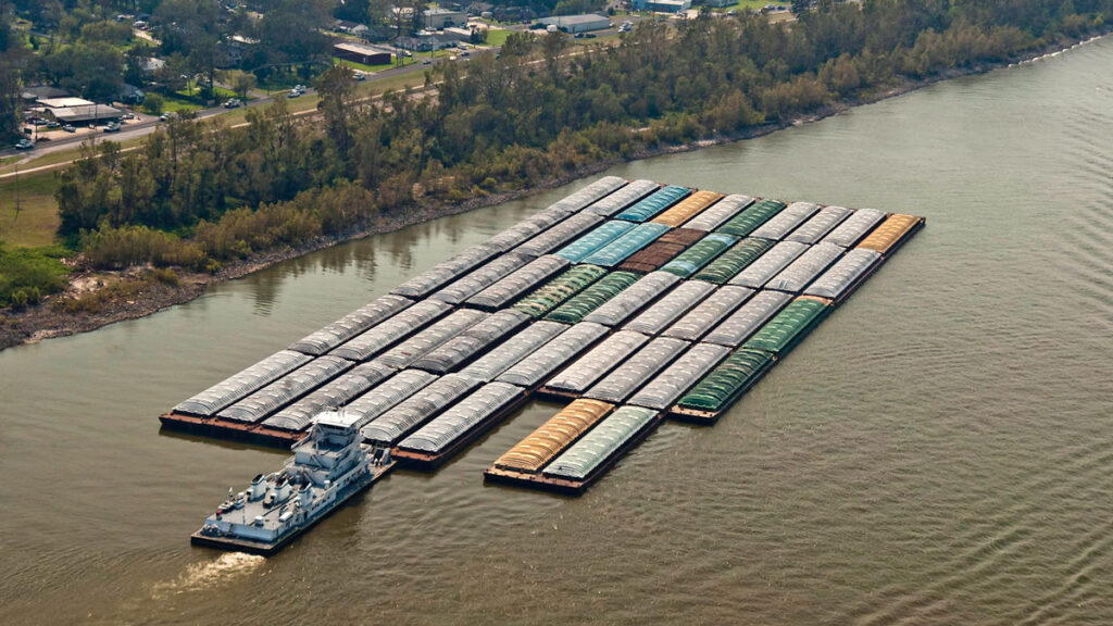 A large boat pushes a raft of dozens of barges down the muddy Mississippi River.