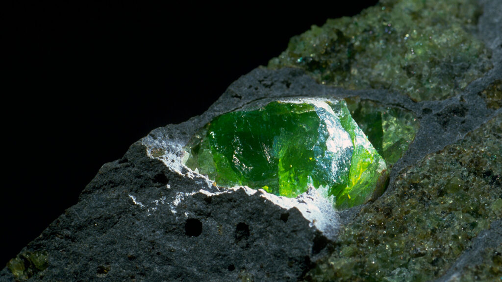 A brilliant green mineral juts out from a chunk of blackish rock dotted with other patches of green crystals.