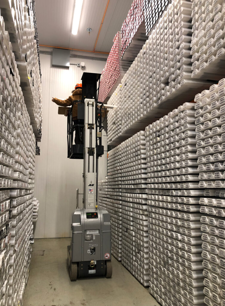A curator stands in a basket atop a movable hydraulic lift, pulling a sediment core from a storage rack.