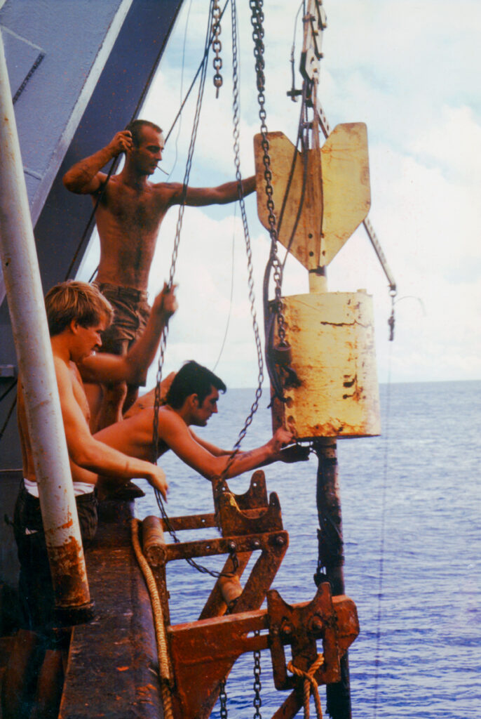 Three people work to deploy a sediment coring device over the side of a ship.