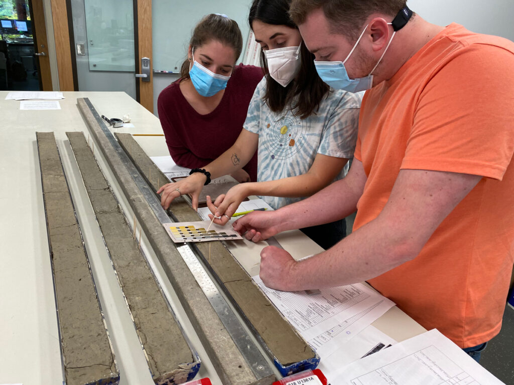 Three people stand beside a table examining sediment cores.