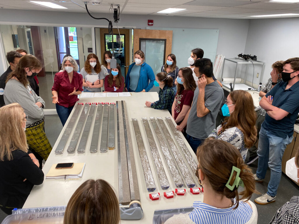 A group of people stand around a table holding several sediment core sections, listening to an instructor speak.