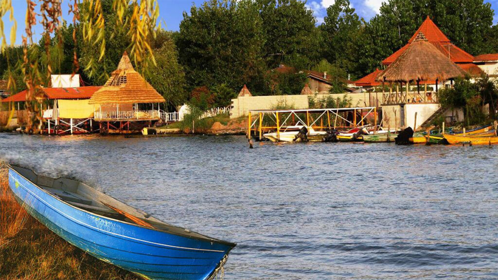 In this photo of a blue lagoon, a small blue boat and the leaves of a weeping willow are in the foreground. On the far side of the water are more boats and several structures with round, pointed roofs.