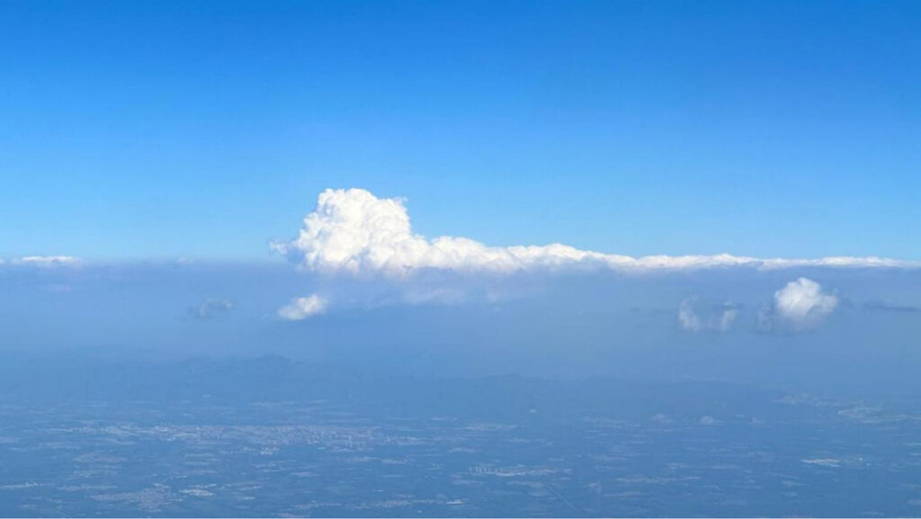 A layer of fluffy white clouds hovers over a hazy landscape, as seen out the window of an airplane.