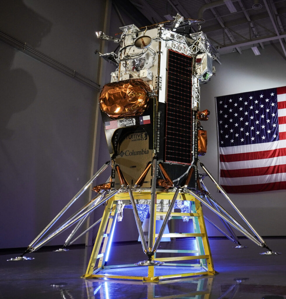 Photograph of a lunar lander in a dimly lit hanger in front of an American flag