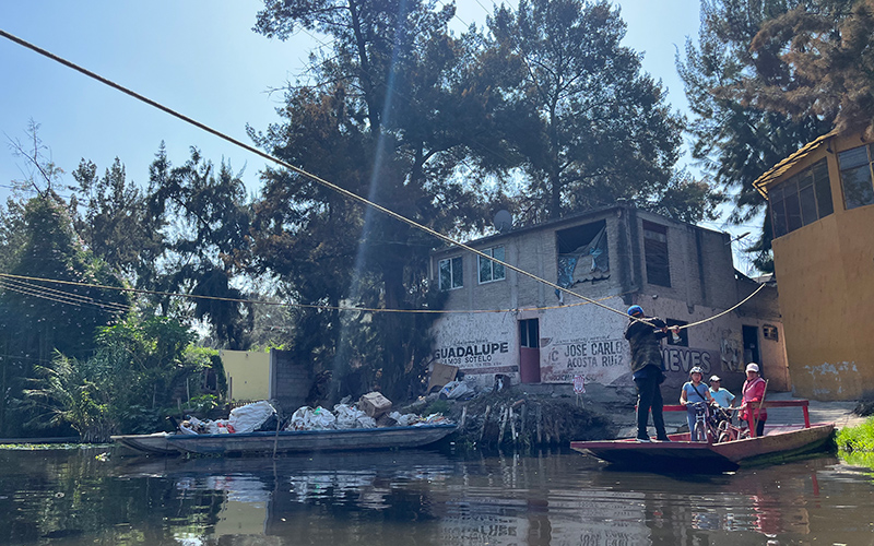 Several people sit on a small boat waiting to be pulled across a waterway in front of houses and near another boat loaded with garbage.