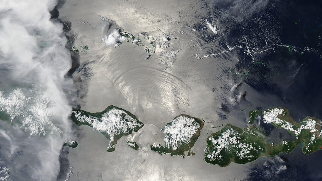 A satellite image of a chain of islands. The ocean looks glossy and mirrorlike, with a large ripple expanding upward from between two of the landmasses.