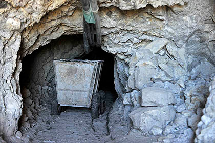 A cart stands in the entrance to a silver mine. The entrance is roughly round and connects to the blackness of a tunnel that extends beyond the field of view. Surrounding the mine entrance are rough-cut rocks. A pile of rock rubble rests to the right of the mine entrance.