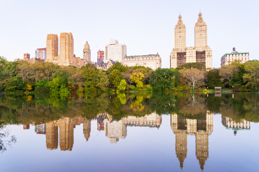 A photo of Central Park in New York City shows a lake in the foreground, trees in the midground, and skyscrapers in the background. The trees and buildings are reflected in the lake.