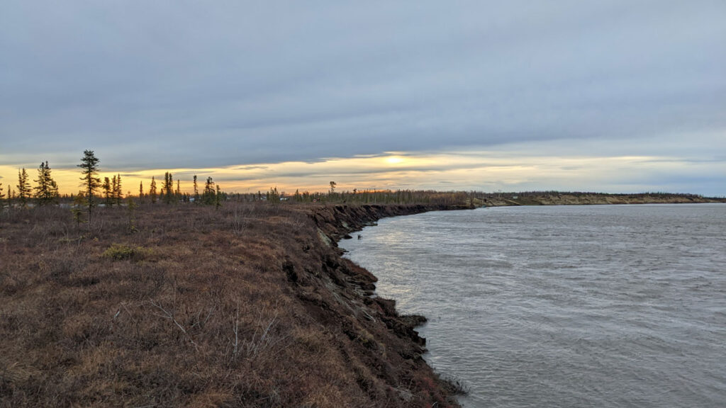 A coastline in Alaska curves off into the distance. The sky is cloudy and the Sun is setting.