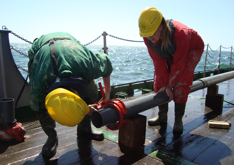 Two people wearing yellow hard hats work with a sediment core onboard R/V Poseidon in 2010.