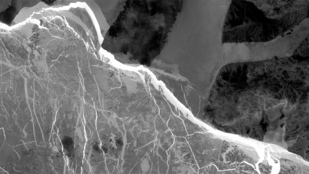 A black and white satellite image shows sea ice, with cracks appearing bright white, beside snow-covered landfast ice (gray) and land (dark).
