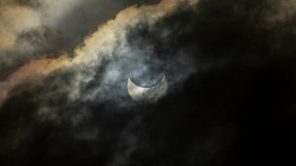 A bright circle of light appears behind clouds, and part of that circle is obscured.