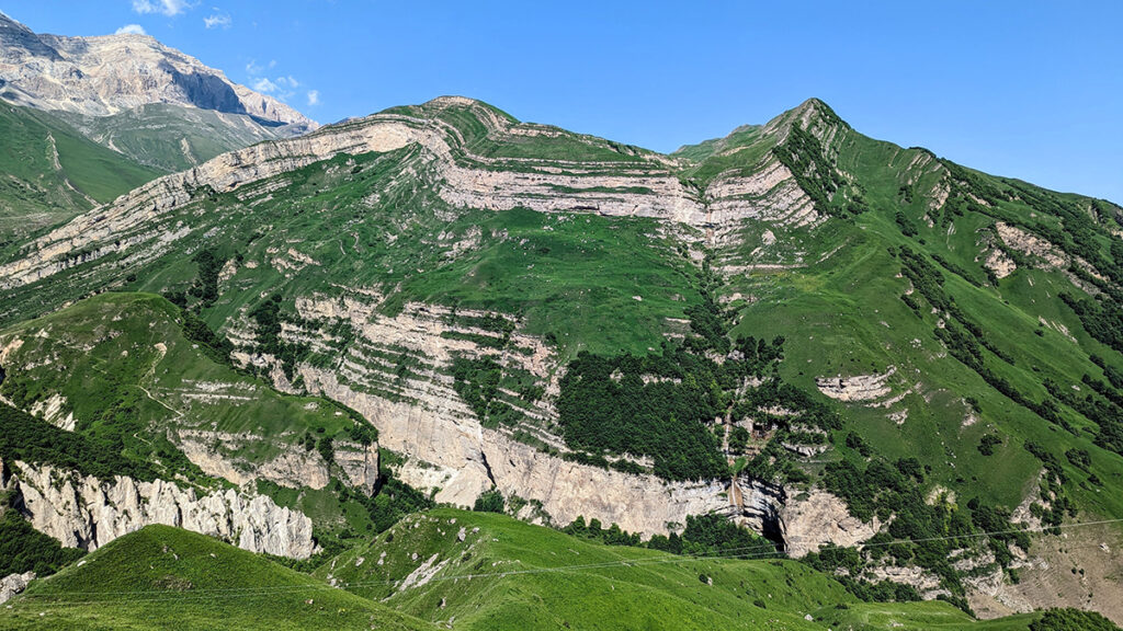 A mountain with alternating stripes of greenery and bare beige rock. There is a point in the middle of the rock where the stripes change direction, indicating a fault propagation fold.