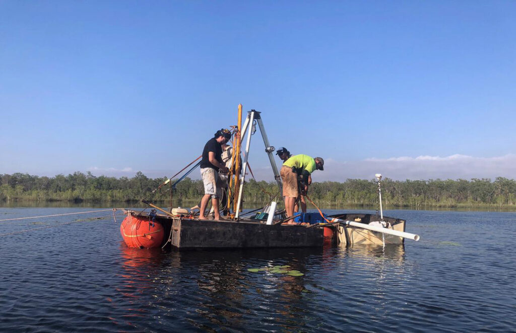 Four people stand on a floating platform on a lagoon. They have a tripod set up and are working