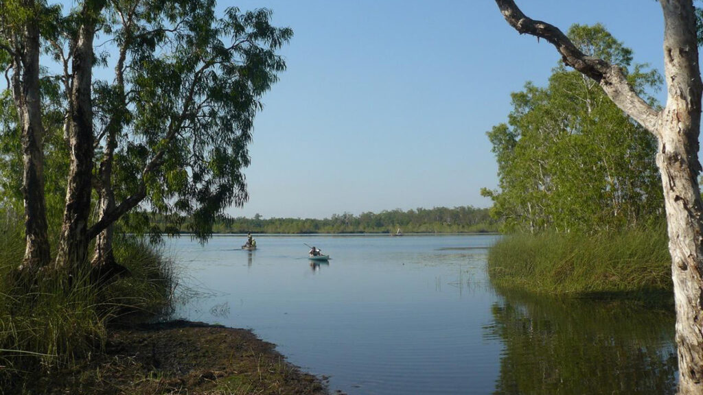 Two people row boats across a blue lagoon, which is flanked by verdant trees.