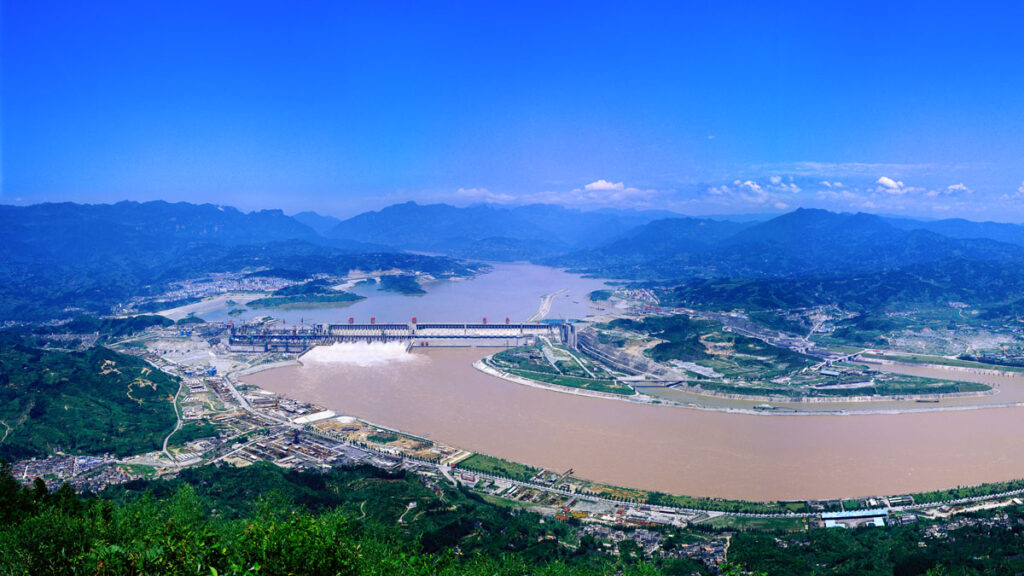 A view of a large river with a dam from above. There are mountains in the distant background and green shrubs in the foreground.