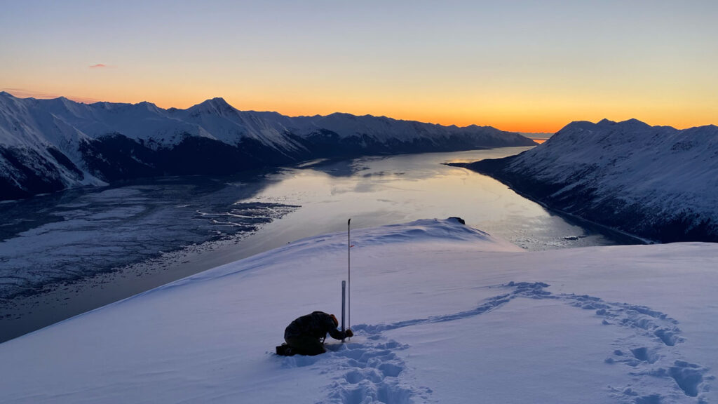 A scientist kneels in the snow with instruments to measure it with a wide view of the Chugach Mountains.