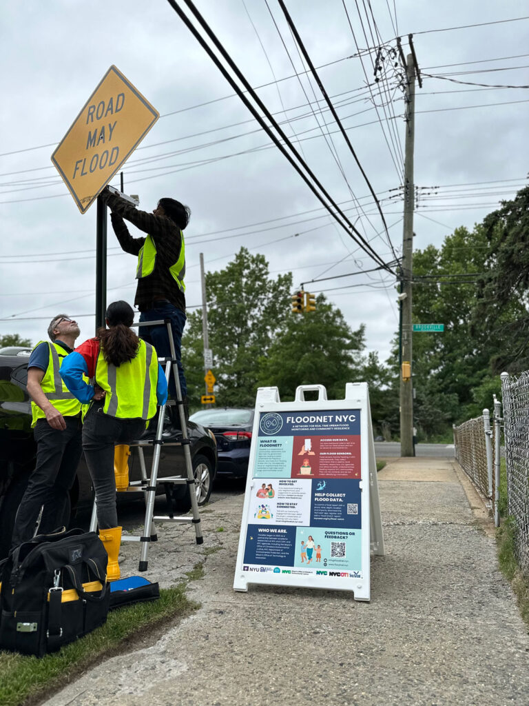 Three people in yellow vests attach a sensor to a street sign that reads “Road may flood.” Next to them is a smaller sign that reads “Floodnet NYC.”