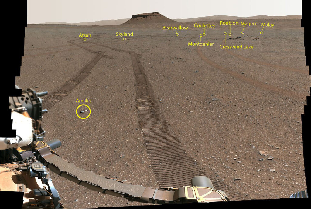 Ten small metal tubes sit on Mars’s rocky orange surface amid rover treads. Part of the rover is visible in the foreground, and a rocky mound sits in the background.