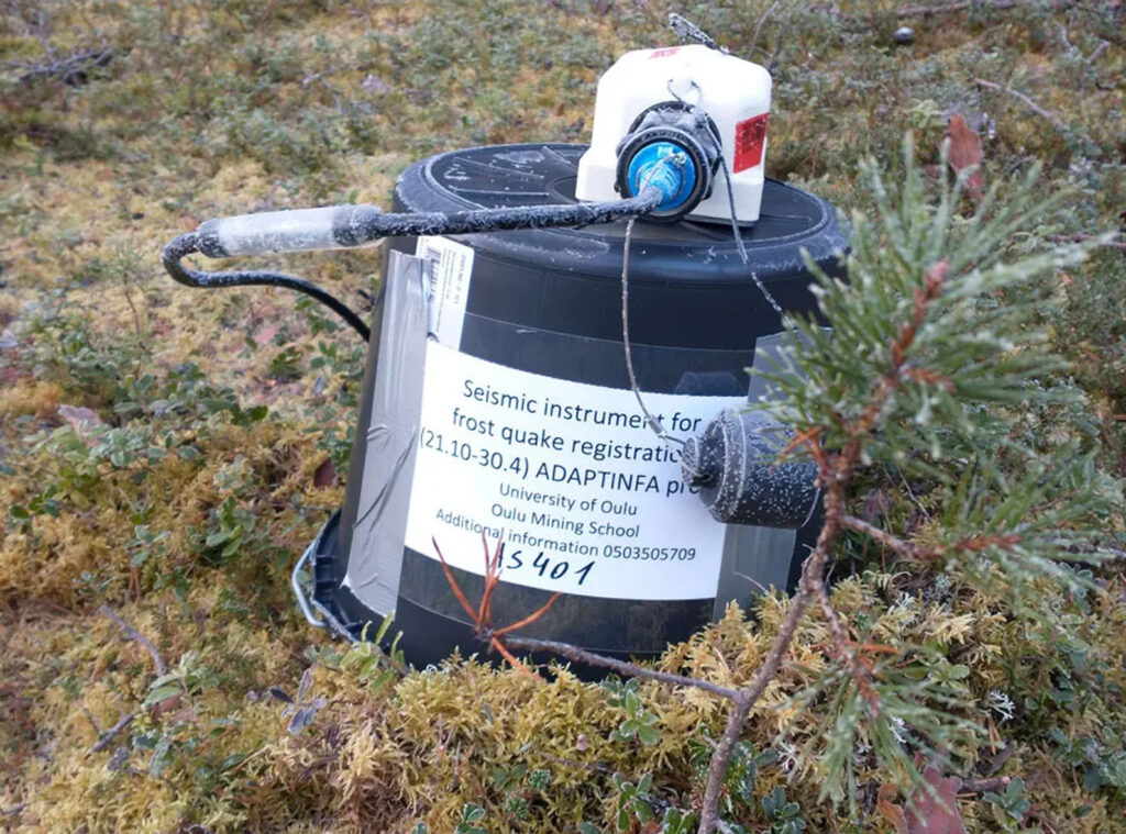 A black, cylindrical seismic instrument sits on a bed of moss covered in a thin layer of frost.