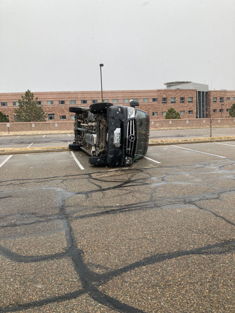 A black Mercedes van blown over by wind sits on its side in a parking lot.