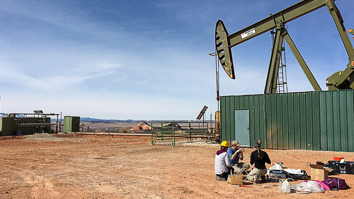 An oil rig surrounded by a green wall sits on a dirt landscape. Three people, surrounded by boxes and bags, sit in the dirt and examine instruments.