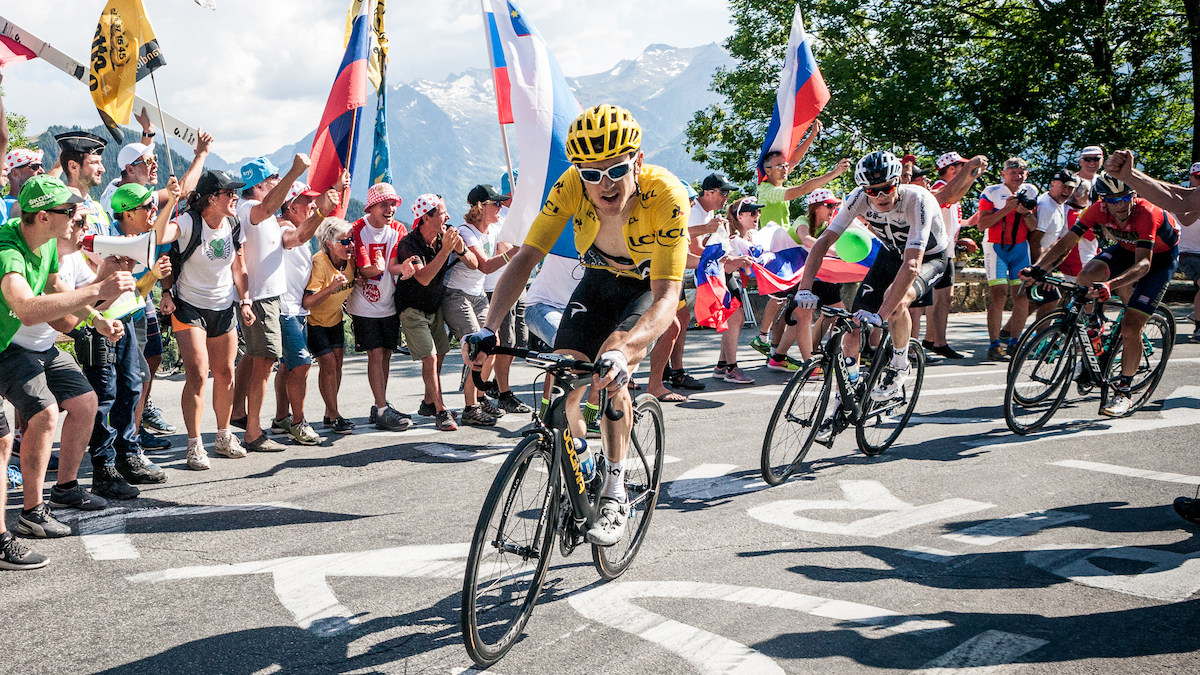 A group of cyclists rounds a corner on a mountain road as a crowd of fans cheers during the 2018 Tour de France.