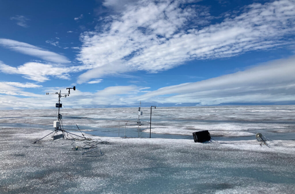 Several large tripods and other weather equipment are set up on a sheet of ice, which stretches into the horizon. The sky is blue and streaked with clouds.