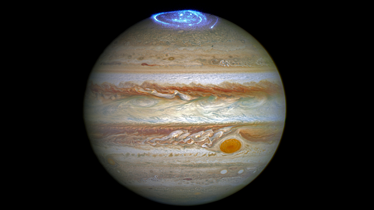 The planet Jupiter, with its stripes and swirls in cream, brown, and orange tones and signature orange dot, is shown against a black background. Bright blue and white aurorae swirl at the top of the planet.