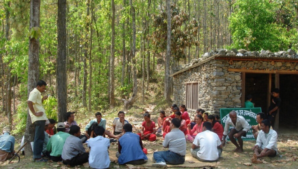 A group of about 30 people, most sitting in a circle on the ground, meet beside a stone building in the forest as part of a community forest user group.
