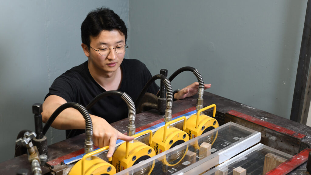 A young man wearing glasses operates an earthquake experiment setup. Four yellow cylinders attached to metallic tubes sit beside a layer of plexiglass held up by wooden columns.