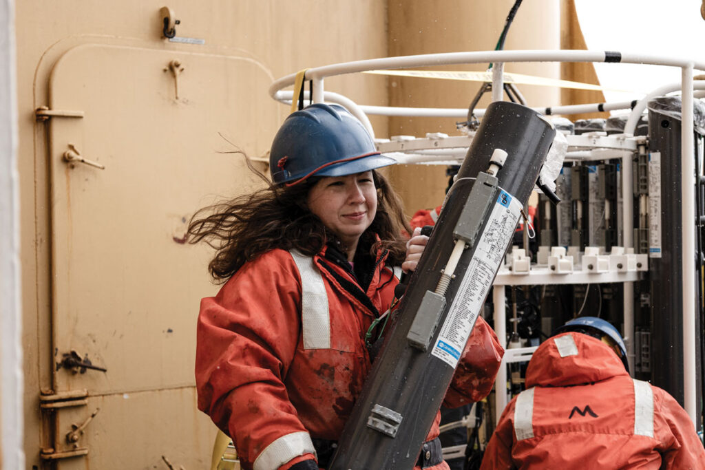 A scientist outfitted in a float coat and hardhat carries a long cylinder.