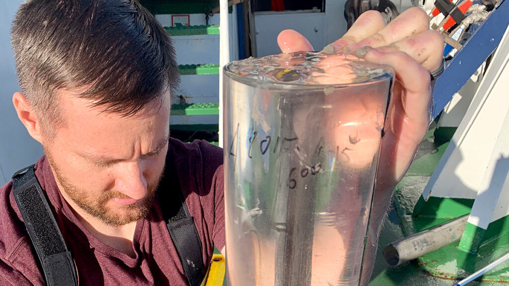 A man holds a clear cylinder that is about 3 feet tall and holds ocean sediment and water.
