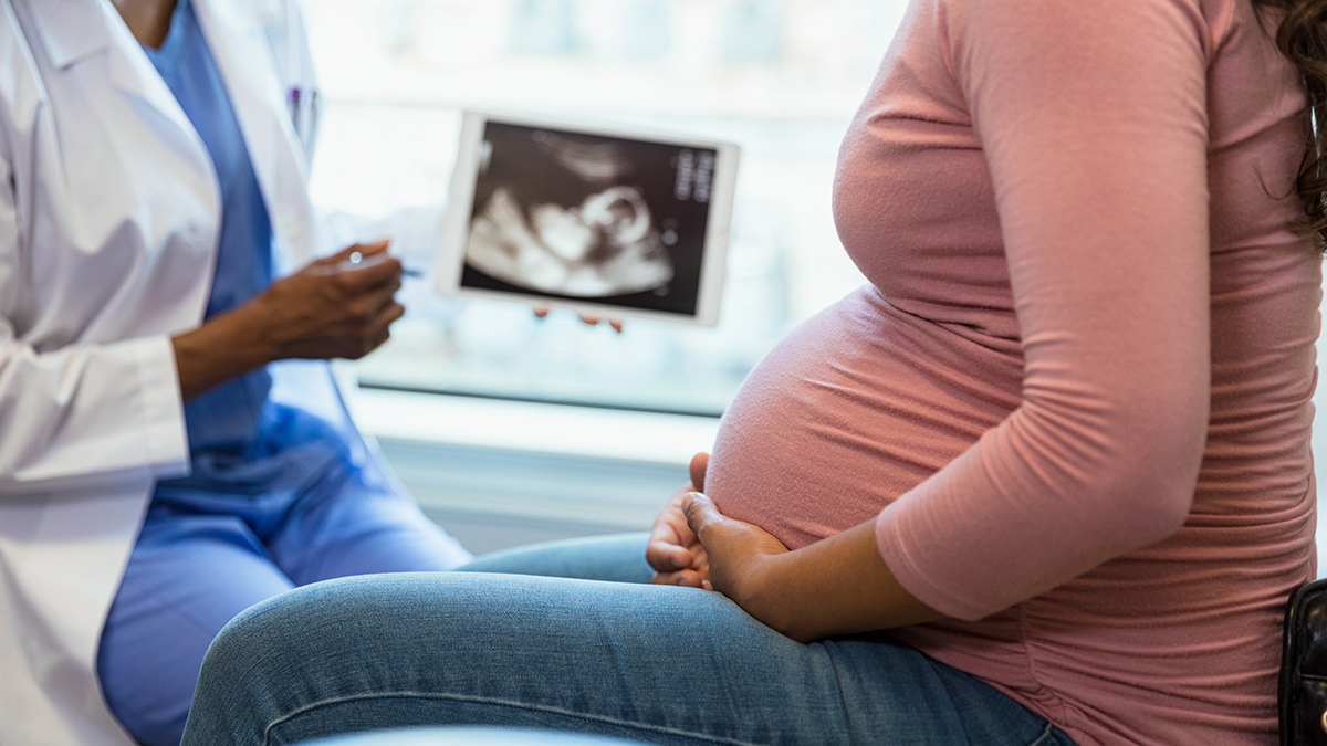 In the foreground at right is an unrecognizable pregnant woman seated in profile, with her hands cradling her abdomen. In the background is an unrecognizable person wearing blue scrubs and a white jacket, holding a sonogram.