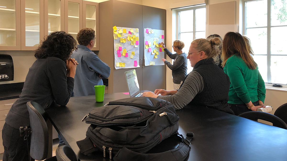 Six people sitting and standing around a black table in a classroom look on as another person writes on a large piece of paper affixed to a cabinet door.