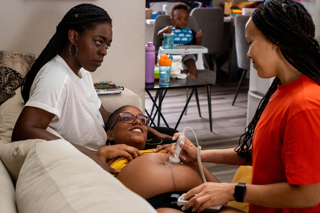 A pregnant woman lies on the couch with her head in her wife’s lap. A midwife is doing a sonogram on her belly, and a toddler sits in a high chair nearby.