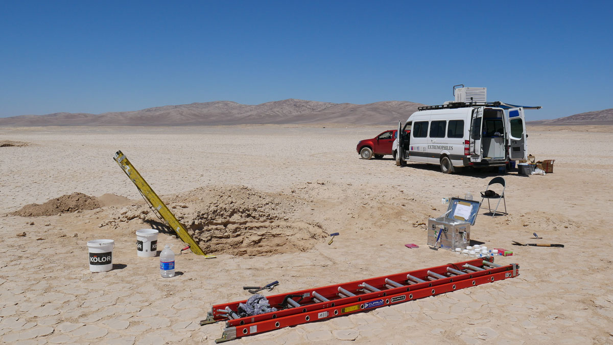 A van labeled “Extremophiles” stands next to ladders extending into a small, deep pit dug into the Atacama Desert.